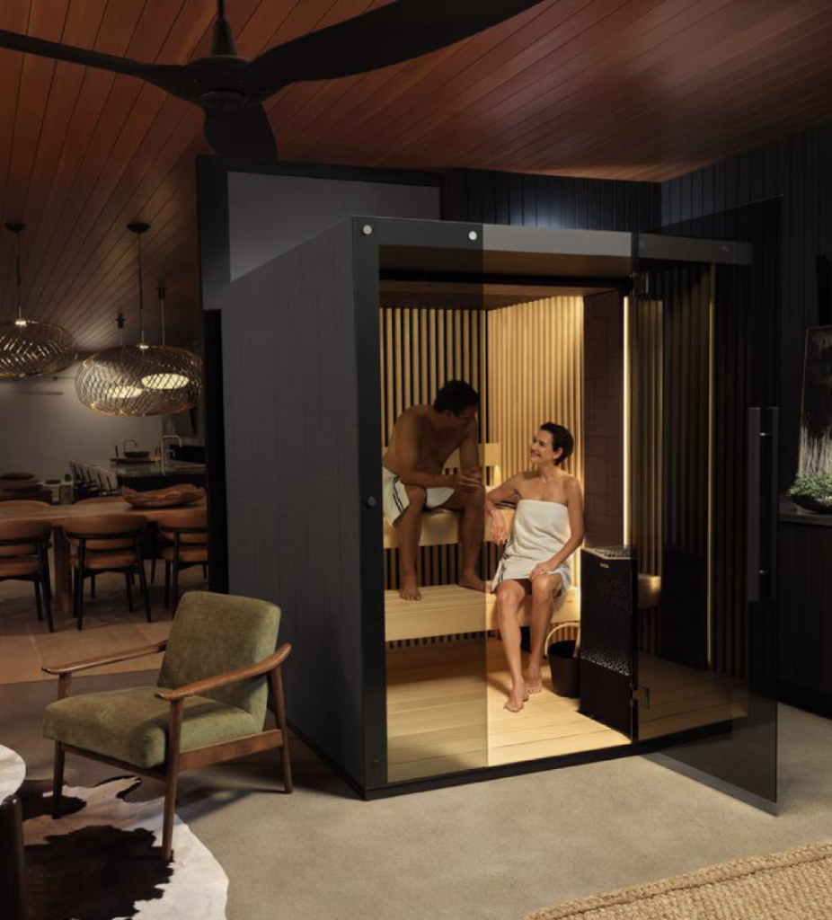 Two people relax in a modern wooden sauna. The room features stylish furniture and warm lighting, creating a cozy, inviting atmosphere.